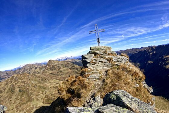 Ein metallisches Gipfelkreuz auf einem felsigen Berggipfel vor blauem Himmel und Gebirgspanorama.