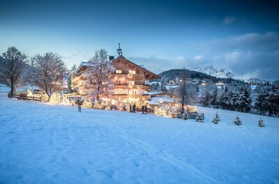 Beleuchtetes Holzhotel in verschneiter Landschaft bei Dämmerung mit geschmückten Tannenbäumen.