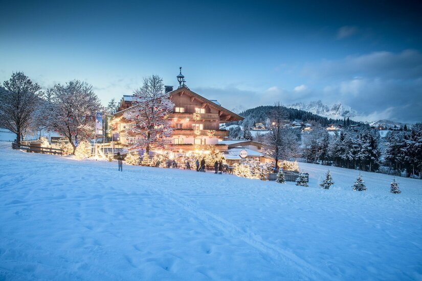 Advent am Rasmushof Kitzbühel Beleuchtetes Holzhotel in verschneiter Landschaft bei Dämmerung mit geschmückten Tannenbäumen.