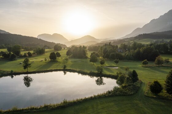 Ellmau_See beim Golfplatz_Wilder Kaiser Eine malerische Landschaft mit sanften Hügeln und einem ruhigen Teich. Die Sonne geht hinter den Bergen unter und taucht die Szene in warmes Licht.