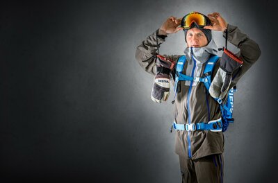 David Kreiner1 Ein Mann trägt eine Skiausrüstung und hält eine Skibrille in den Händen. Er steht vor einem grauen Hintergrund und wirkt bereit für ein Abenteuer im Schnee.