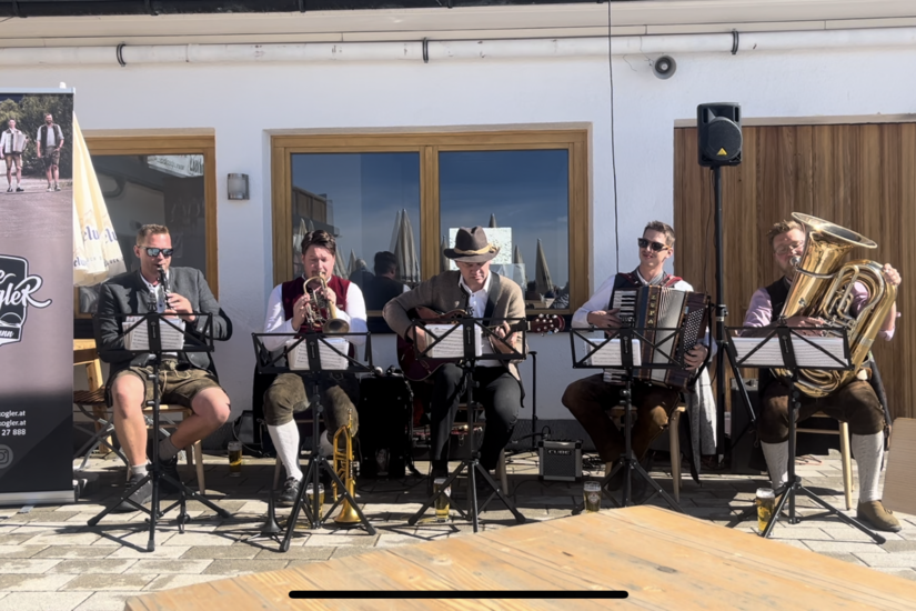 Fünf Musiker in Tracht spielen Blasinstrumente, Gitarre und Akkordeon auf einer Terrasse vor einem Gebäude.