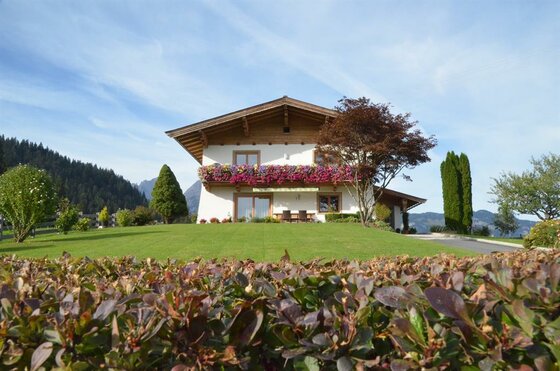 Haus Jöchl in Reith Zweistöckiges Alpenhaus mit Blumenbalkon und gepflegtem Rasen vor bewaldeten Bergen.