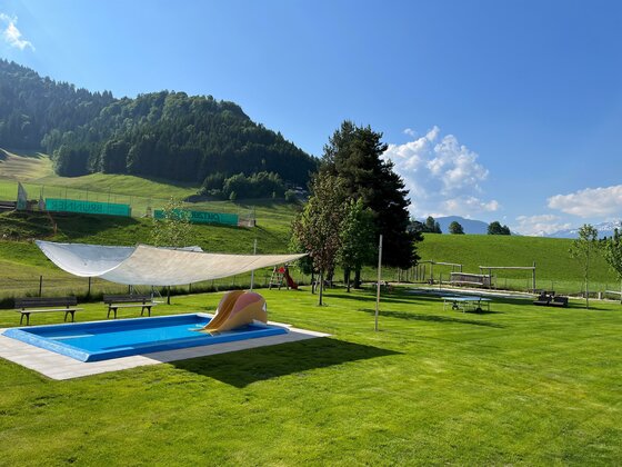 Ein schöner Garten mit einem blauen Pool und einer Liegewiese. Im Hintergrund sind Hügel und ein klarer Himmel zu sehen.