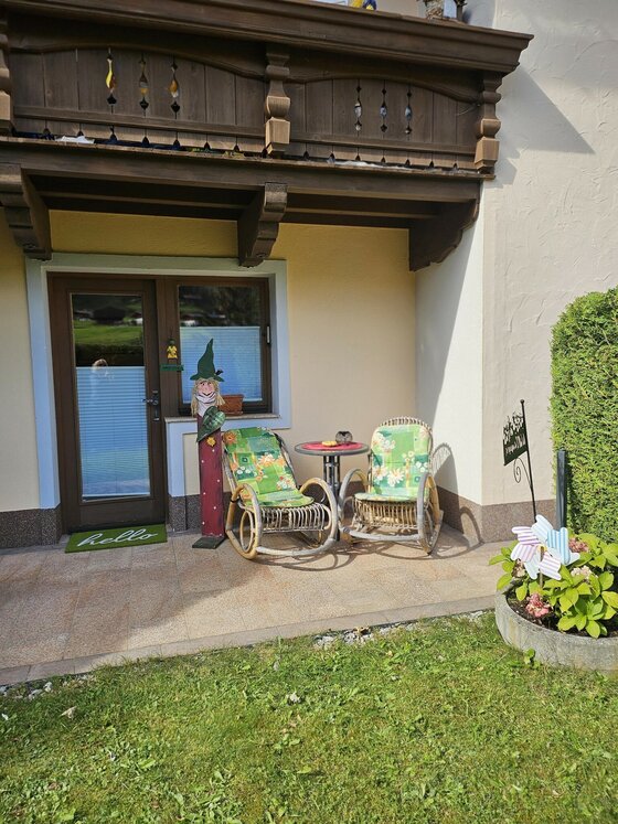Terrasse bei Tamara's Apart Zwei Korbsessel mit grünen Polstern und ein kleiner Tisch auf einer gepflasterten Terrasse unter einem Holzbalkon.