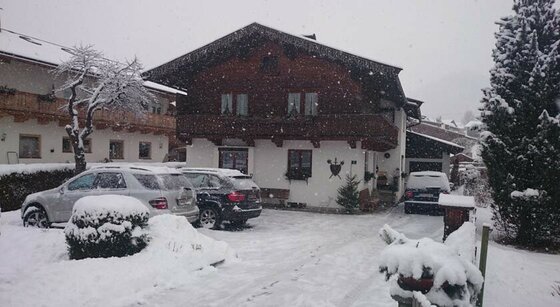 Reason Winter Außenansicht Alpines Wohnhaus mit Holzfassade und Balkon bei Schneefall mit parkenden Autos davor.