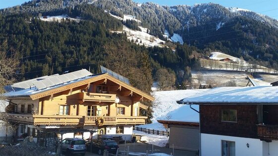 Alpines Holzhaus mit Solaranlage vor bewaldetem Berghang im Winter.