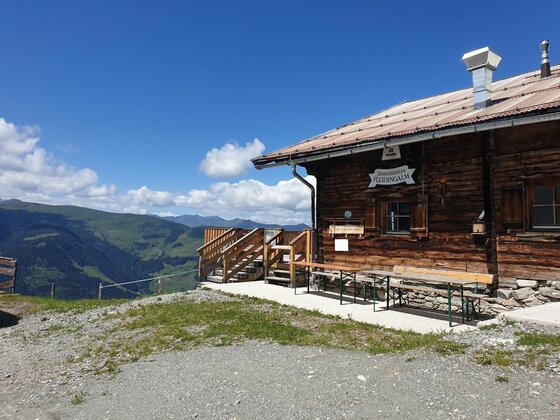 Rustikale Holzhütte mit Sitzbänken vor Gebirgspanorama unter blauem Himmel.
