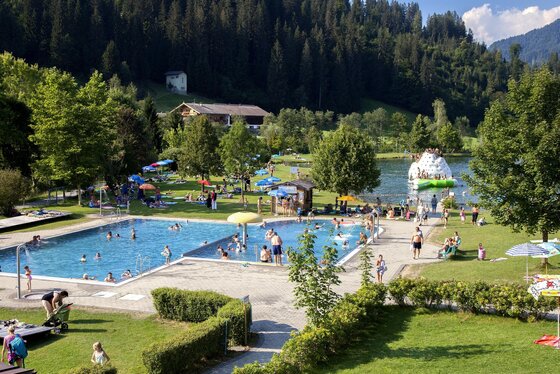 Ein wunderschöner Schwimmbereich mit zwei Becken und viel Grün. Menschen genießen die Sonne und das Wasser in einer ruhigen, naturnahen Umgebung.