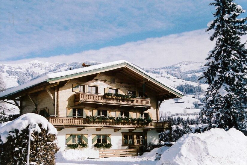 Alpines Holzhaus mit Balkonen im Schnee vor bewaldeten Bergen.