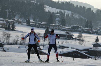 Zwei Personen beim Skilanglauf auf einer verschneiten Loipe vor einer dörflichen Kulisse.