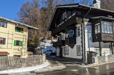Zwei mehrstöckige Häuser im alpinen Stil an einer asphaltierten Straße mit Schneeresten unter blauem Himmel.