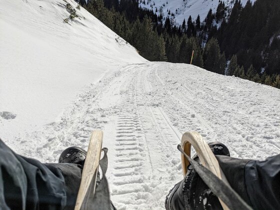 Rodeln bei der Kelchalm Blick von einem Holzschlitten auf eine präparierte Schneepiste im Gebirge mit Nadelwald im Hintergrund.
