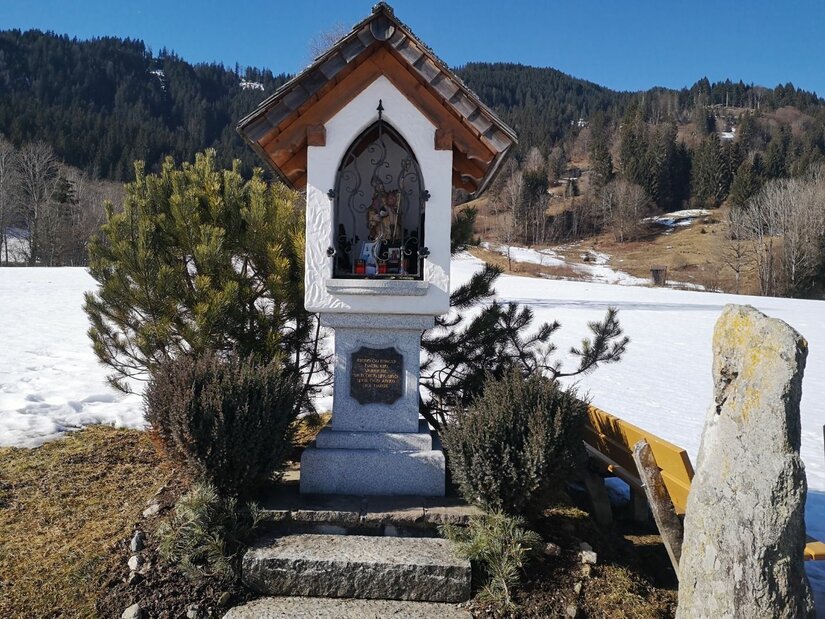 Ein weißer Bildstock mit Satteldach vor einer verschneiten Berglandschaft.