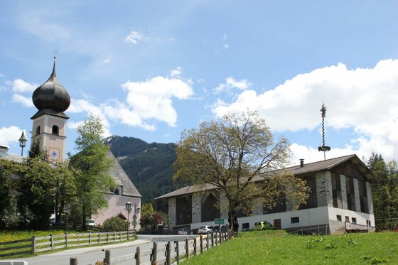 Ortsansicht mit Kirche Kirche mit Zwiebelturm und ein größeres Holzgebäude neben einer Straße vor bewaldeten Bergen.