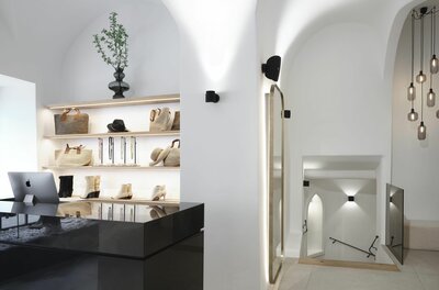 Moderner Verkaufsraum mit weißen Gewölbewänden, beleuchteten Regalen für Taschen und Schuhe sowie einem Treppenabgang.