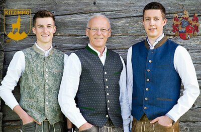 Drei Männer stehen vor einer Holzwand und tragen traditionelle Trachtenwesten. Sie lächeln freundlich in die Kamera.