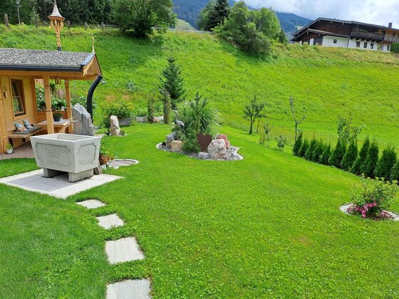 Garten der Ferienwohnung Widmann Gartenanlage mit Holzhütte, Steintrog und gepflegter Rasenfläche vor einem grünen Hang.