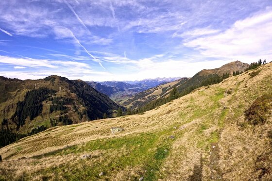 Aussicht auf der Schotting-Runde Blick von einem grasbewachsenen Berghang in ein weites Tal unter blauem Himmel mit Kondensstreifen.