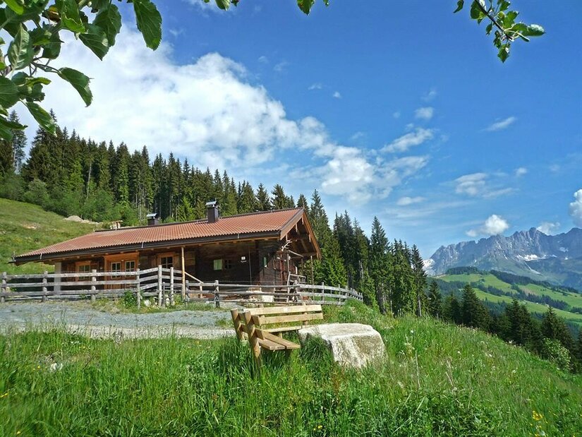Holzhütte mit Zaun auf einer grünen Wiese vor Wald und Gebirgskette unter blauem Himmel.