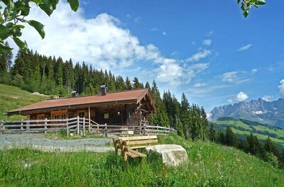 Holzhütte mit Zaun auf einer grünen Wiese vor Wald und Gebirgskette unter blauem Himmel.