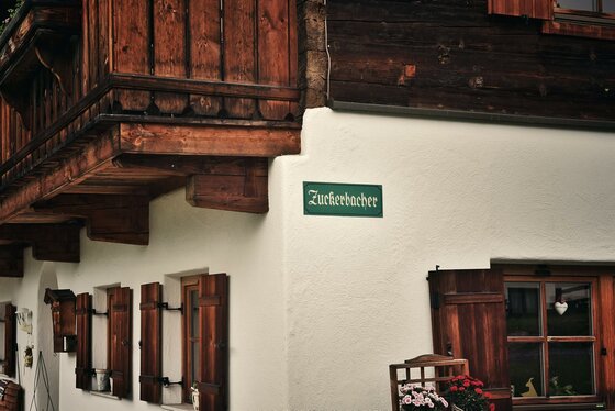 Nahaufnahme einer Hauswand mit Holzfensterläden, Balkon und einem grünen Schild mit der Aufschrift Zuckerbacher.