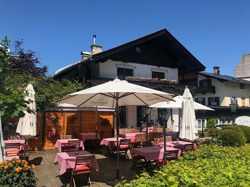 Außenterrasse eines Restaurants mit rot-weiß karierten Tischdecken und weißen Sonnenschirmen vor einem Alpenhaus.