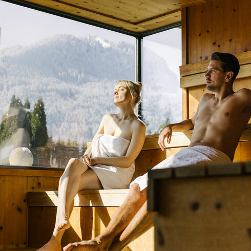 Ein Paar entspannt in einer modernen Sauna mit Holzverkleidung, während die Sonne durch große Fenster scheint.
