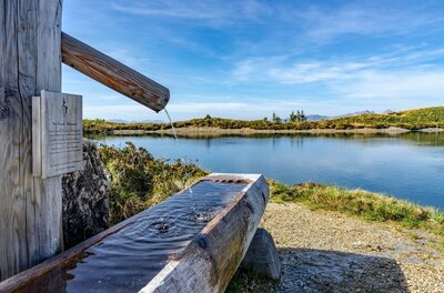 Holzbrunnen mit fließendem Wasser vor einem Bergsee unter blauem Himmel.