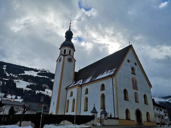 Pfarrkirche vor Bergkulisse Weiße Kirche mit Zwiebelturm vor bewaldeten, schneebedeckten Bergen unter bewölktem Himmel.