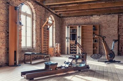 Fitnessraum mit Holzgeräten, Rudermaschine und Ergometer vor einer Ziegelwand.