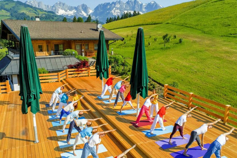 Sivananda Yoga Eine Yoga-Gruppe praktiziert im Freien auf einer Holzterrasse mit Blick auf die Berge. Der Himmel ist klar und es ist sonnig.