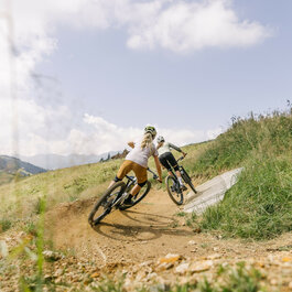 Sonnenrast Trail in Kitzbühel, zwei Mountainbiker fahren durch Kurven auf einem grünen Trail unter blauem Himmel.
