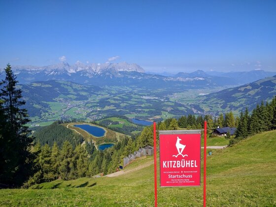 Panoramablick am Hahnenkamm Blick vom Hahnenkamm auf Kitzbühel mit rotem Startschuss-Schild, Speicherseen und Gebirgskette im Hintergrund.
