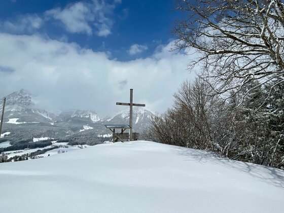Holzkreuz auf einem verschneiten Hügel vor einem Bergpanorama unter blauem Himmel.
