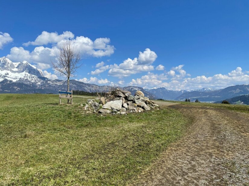 Ein Steinhaufen neben einem Feldweg auf einer Wiese vor schneebedeckten Bergen unter blauem Himmel.