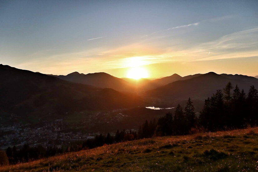 Sonnenuntergang über einer bergigen Landschaft mit einem Tal und Nadelbäumen im Vordergrund.