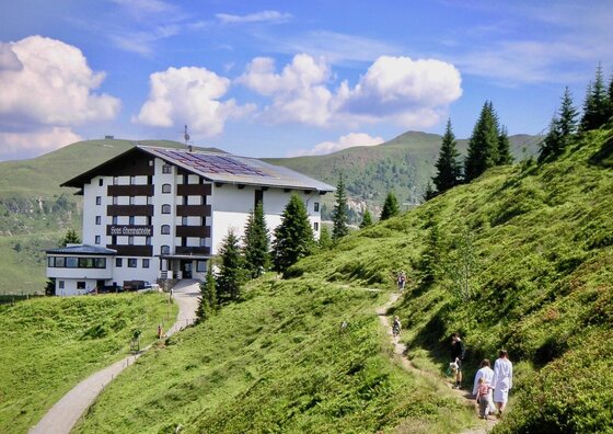 Berghotel Ehrenbachhöhe im Sommer Mehrstöckiges Berghotel an einem grünen Hang mit Wanderern auf einem schmalen Pfad im Vordergrund.