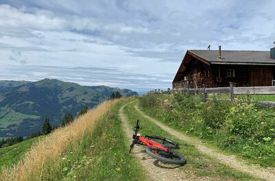 Ein rotes Mountainbike liegt auf einem Schotterweg neben einer Holzhütte vor Bergpanorama.
