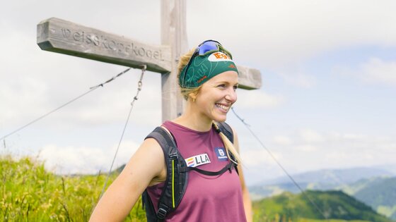 Eine Wanderin mit Stirnband und Rucksack steht lächelnd vor dem hölzernen Gipfelkreuz des Weißkopfkogels.