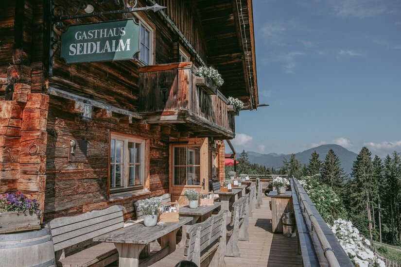 Gasthaus Seidlalm Terrasse Holzfassade des Gasthauses Seidlalm mit Außenterrasse, Holztischen und Blick auf bewaldete Berge.