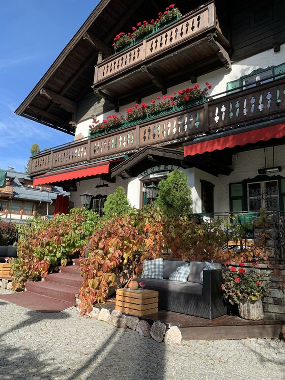 Außenansicht Villa Licht Alpines Hotelgebäude mit Holzbalkonen, roten Markisen und einer Terrasse mit Sitzmöbeln.