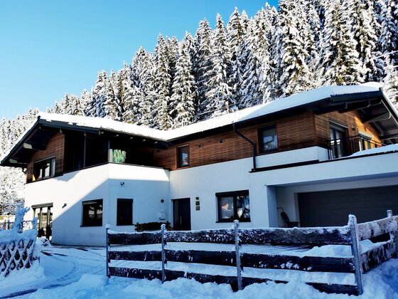 Modernes zweistöckiges Haus mit Holzfassade und schneebedecktem Dach vor einem verschneiten Nadelwald.