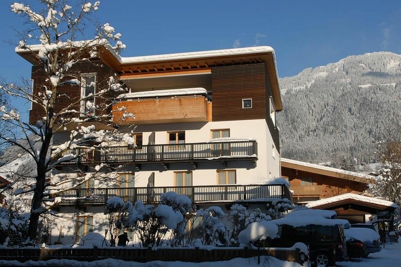 Mehrstöckiges Gästehaus mit Holzbalkonen im Winter vor verschneitem Berghang.