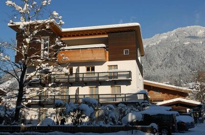 Mehrstöckiges Gästehaus mit Holzbalkonen im Winter vor verschneitem Berghang.
