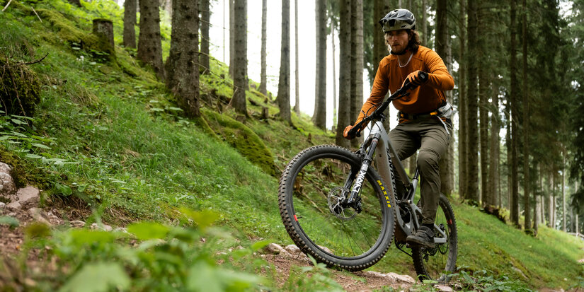 In Kitzbühel fährt ein Radfahrer in orangefarbenem Oberteil sicher über steinigen Weg durch dichten Wald.