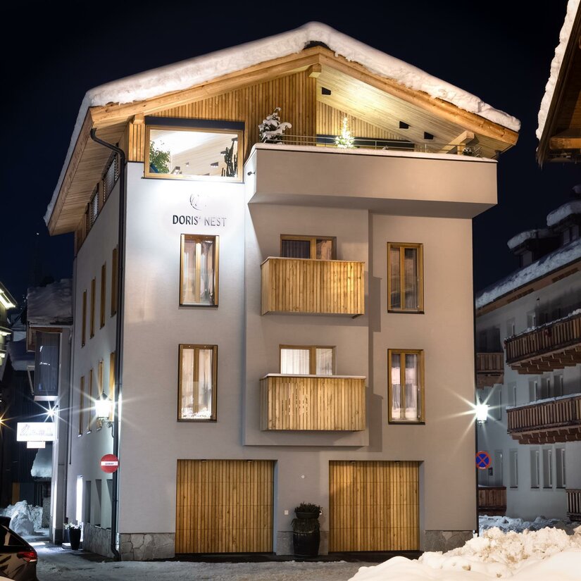 Beleuchtetes, mehrstöckiges Gebäude mit Holzfassadenelementen und schneebedecktem Dach bei Nacht.