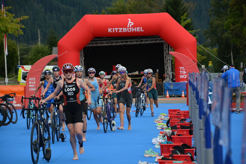 Athleten in Triathlon-Outfits laufen mit Fahrrädern über einen blauen Teppich, während Zuschauer im Hintergrund zuschauen.