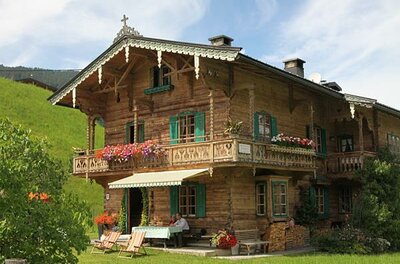 Zweistöckiges Holzhaus im alpinen Stil mit Blumenbalkonen und grünen Fensterläden vor einer Wiese.