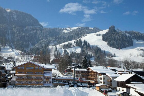 Kaiserhof Winter in Kitzbühel Verschneite Hotelanlage am Fuße eines bewaldeten Berges mit Skipisten und Seilbahn unter blauem Himmel.